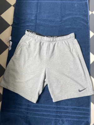 Grå Nike shorts i bomull - Snygga ljusgrå shorts från Nike med elastisk midja och diskret logga på benet. Tillverkade i mjuk bomull för maximal komfort. Perfekta för sommardagar eller träning. Klassisk passform och stilren design.