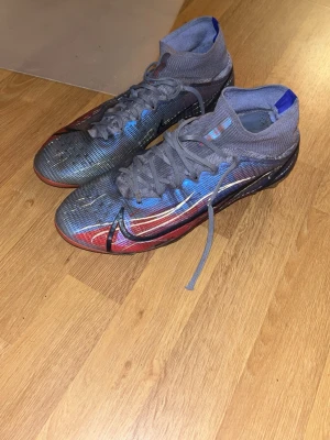 Nike Mercurial Superfly fotbollsskor - Säljer ett par Nike Mercurial Superfly fotbollsskor med högt skaft. Skorna har en grå bas med blå, röda och vita detaljer samt den klassiska Nike-swooshen på sidan. Ovandelen är i syntetmaterial för bra bollkontroll och passform. Perfekta för dig som vill sticka ut på planen.