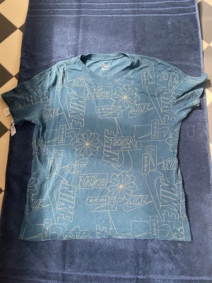 Blå Nike t-shirt med grafiskt tryck - Säljer en blå t-shirt från Nike med coolt grafiskt tryck över hela plagget. T-shirten har korta ärmar och rund halsringning. Trycket består av Nike-loggor, blommor och texten 'Just do it' i vitt. Perfekt för en avslappnad och sportig stil. Den är i ett bra skick, den enda defekten är texten där storleken står (Sista bilden).