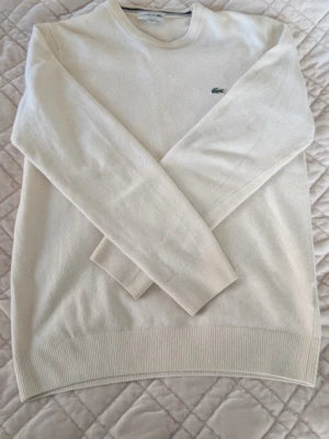 Beige stickad tröja från Lacoste - Snygg beige stickad tröja från Lacoste med klassisk rund halsringning och den ikoniska gröna krokodilloggan på bröstet. Tröjan har ribbade muddar vid ärmslut och nederkant. Perfekt för dig som gillar stilrena och tidlösa plagg.