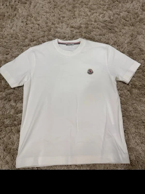 Vit t-shirt från Moncler - Klassisk vit t-shirt från Moncler med rund halsringning och korta ärmar. T-shirten har en liten Moncler-logga broderad på bröstet och är tillverkad i mjuk bomull för en skön känsla. Perfekt basplagg med stilren design.