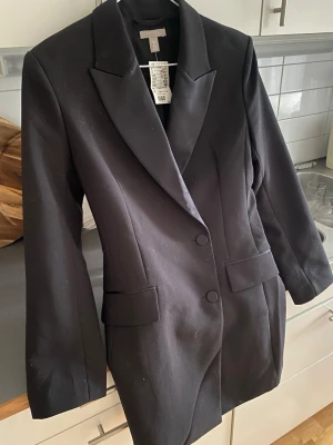 Svart kavaj från H&M - Stilren svart kavaj från H&M  och två knappar framtill. Kavajen har två fickor med lock och en figurnära passform som ger en snygg siluett. Perfekt för dig som vill ha en klassisk och trendig look. Ny