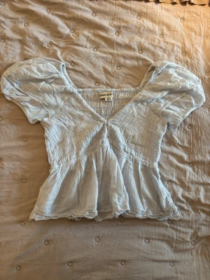 Ljusblå blus från Urban Outfitters - Supersöt ljusblå blus från Urban Outfitters med puffiga korta ärmar och v-ringning. Toppen har smockdetaljer över hela och en lätt volang nertill. Perfekt för dig som gillar romantisk och luftig stil. Liten fläck finns på armen men det syns inte allt för mycket 