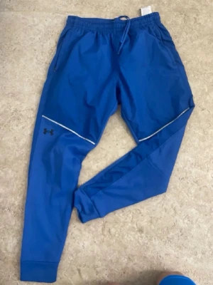Blå träningsbyxor från Under Armour - Snygga blå träningsbyxor från Under Armour med resår och dragsko i midjan. Byxorna har dragkedjefickor, reflexdetaljer och smal passform med muddar vid bensluten. Perfekta för löpning eller gym.