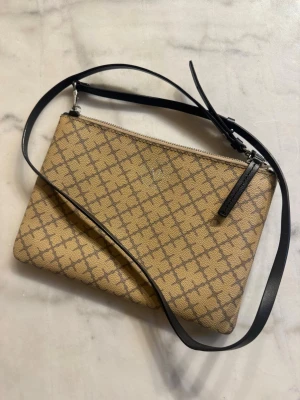 Beige axelväska från By Malene Birger - Snygg beige axelväska från By Malene Birger med klassiskt monogrammönster i brunt. Väskan har en avlång, rektangulär form och justerbar svart axelrem i läder. Stängs med dragkedja upptill och har en stilren look som passar till det mesta.