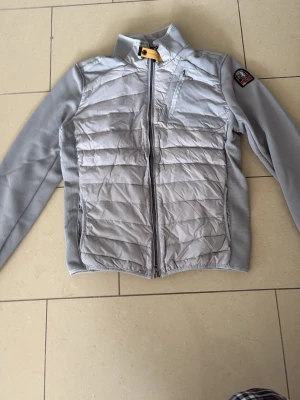 Ljusgrå dunjacka från Parajumpers - Snygg ljusgrå dunjacka från Parajumpers med quiltad front, hög krage och dragkedja. Jackan har en bröstficka med dragkedja och ett patch på ärmen. Materialet är en mix av dun och syntet, perfekt för kalla dagar.