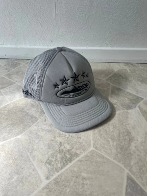 Grå meshkeps från Ciele Athletics - Snygg grå keps från Ciele Athletics med meshpaneler och justerbar snapback. Framtill finns broderade svarta stjärnor och logga, samt broderad text bak. Perfekt för en sportig och avslappnad stil.
