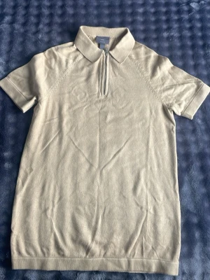 Beige polotopp med dragkedja H&M - Snygg beige polotopp från H&M med korta ärmar och krage. Toppen har en stilren dragkedja framtill som ger en sportig touch. Tillverkad i mjukt bomullsmaterial och passar perfekt till både jeans och kjol.
