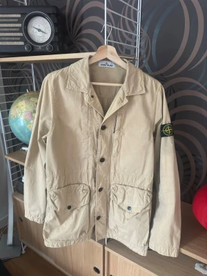 Beige overshirt  Stone Island - Snygg beige overshirt från Stone Island med klassisk patch på ärmen. Jackan har knappar framtill, två stora fickor och en avslappnad krage. Perfekt för lager på lager och streetwear-vibe. Jackan är i väldigt bra skick bara använd få gånger självklart så är den äkta dessutom!