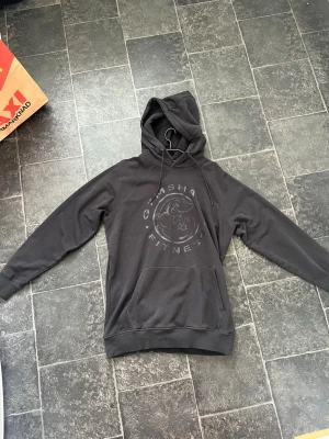 Svart hoodie Gymshark Legacy - Snygg svart hoodie från Gymshark med stort tryck på bröstet som visar en muskulös haj och texten 'Gymshark Fitness'. Hoodien har huva med dragsko och en stor magficka. Perfekt för gymmet eller chill. Mjuk bomullsblandning och avslappnad passform.