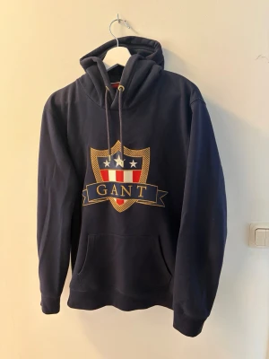 GANT hoodie - Snygg marinblå hoodie från GANT med stort broderat emblem på bröstet. Väldigt skönt material. Verkligen i nyskick, knappt använd alls. Storlek L, sitter TTS.
