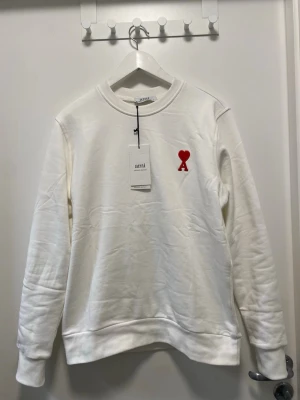 Vit sweatshirt från Ami Paris - Vit sweatshirt från Ami Paris med röd hjärtlogga på bröstet. Klassisk rund halsringning och ribbade muddar vid ärmslut och nederkant. Tillverkad i mjukt material för en clean och stilren look. Passar perfekt till sommaren och till nästan alla outfits. Helt ny med prislapp. Kom med ett prisförslag om du är sugen, eller köp direkt såklart 😉 skickar samma dag!