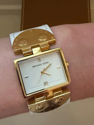 Michael Kors klocka äkta skinn / guld – ny & oanvänd  - Mycket elegant Michael Kors damklocka MK-4053 – helt oanvänd, vit läderrem, gulddetaljer komplett förpackning.  Rektangulär urtavla i vitt med guldtonade visare och datumfönster. Brett vitt äkta läderarmband med snygga guldtonade ståldetaljer — karakteristiska MK-logotypknappar och rörliga ändstycken.  Klockan är aldrig buren och är i perfekt skick utan repor eller märken på vare sig armband eller boett.  Nytt batteri insatt juni 2026.  Märke: Michael Kors Modell: MK-4053 Rörelse: Kvarts med dat