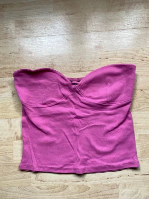 Rosa ribbad bandeau tubtopp - Säljer en rosa bandeau tubtopp i ribbad bomull. Toppen är axelbandslös och har en hjärtformad urringning framtill. Perfekt för varma dagar och enkel att styla med jeans eller kjol.