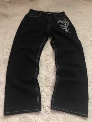 Svarta jeans med vit brodyr - Säljer ett par svarta jeans med raka ben och vit kontrastsöm. På ena benet finns en stor, stilren brodyr i vitt med ett grafiskt motiv. Jeansen har klassisk femficksmodell och är perfekta för dig som vill sticka ut lite extra.