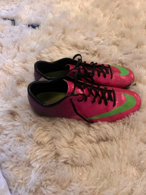Nike färgglada fotbollsskor strl 41 - Säljer ett par retro Nike fotbollsskor i storlek 41 med en riktigt färgstark design. Skorna har en mix av lila, rosa och grönt med svarta snören och en stor Nike-logga på sidan. Yttermaterialet är syntetiskt och sulan är anpassad för spel på gräs.