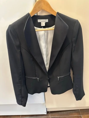 Svart kavaj från H&M med dragkedjor - Stilren svart kavaj från H&M med figurnära passform och två dragkedjefickor framtill. Kavajen har klassisk krage, hakstängning och ljus innerfoder. Perfekt för en trendig och snygg look.