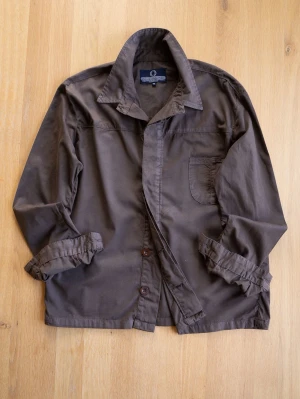 Brun overshirt från Human Scales - Stilren brun overshirt i mjuk bomull. Klassisk krage, knappstängning framtill och en bröstficka. Perfekt lager-på-lager-plagg med avslappnad passform och uppvikta ärmar för en casual look.