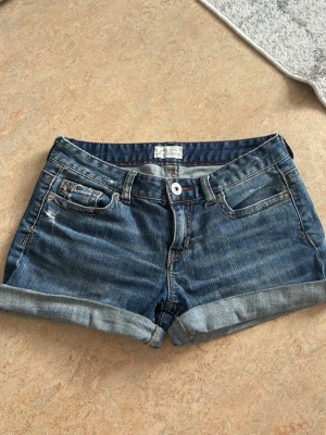 Blå jeansshorts med uppvikta ben - Snygga blå jeansshorts med uppvikta ben och klassiska fem fickor. Shortsen har slitna detaljer och kontrastsömmar, samt bälteshällor och knappgylf. Perfekta för varma dagar och enkel att matcha med olika toppar. Perfekt för dig som gillar lite Y2K style, fint skick, lite svårare att hitta nu då de är en äldre modell och det är et par byxor som klippts till shorts