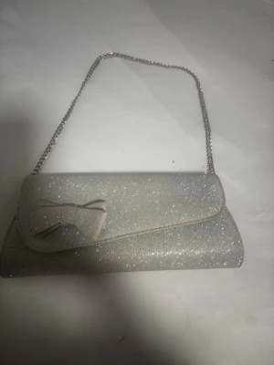 Glittrig silver kuvertväska med rosett - Snygg kuvertväska i glittrigt silvertyg med en dekorativ rosett på locket och en tunn kedjerem. Perfekt för att lyfta din outfit med lite extra bling. Väskan har en klassisk avlång form och stängs med lock. Materialet är tyg med glittereffekt. Den har tappat lite färg 