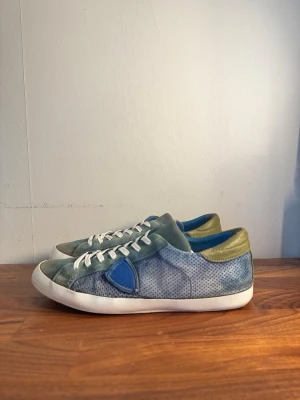 Philippe Model Skor - Snygga sneakers från Philippe Model i blått och grönt mocka och läder. Skorna har vita snören, perforerade detaljer på sidorna och en blå logotyp-patch. Insidan är blå och hälen har en olivgrön detalj. Perfekt för dig som gillar unika färgkombinationer.
