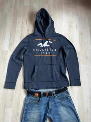 Mörkblå hoodie från Hollister - Mörkblå hoodie från Hollister med stor vit logga och texten 'HOLLISTER 1922' framtill. Hoodien har huva med dragsko, magficka och orangea detaljer. Tillverkad i mjukt bomullsmaterial och har långa ärmar. Perfekt för en avslappnad och cool stil. Storlek S