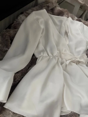Vit omlottblus i satin - Snygg vit omlottblus i glansigt satinmaterial med knytband i midjan och långa ärmar. Perfekt för dig som gillar en stilren och elegant look. Blusen har en lätt figursydd passform och känns lyxig mot huden.