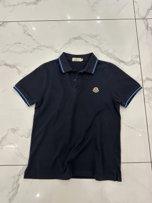 Marinblå piké från Moncler - Marinblå pikétröja från Moncler i bomull med klassisk krage och blå/vita ränder på krage och ärmslut. Kortärmad modell med knappslå och Moncler-logga på bröstet. Enkel och stilren design som funkar till det mesta.