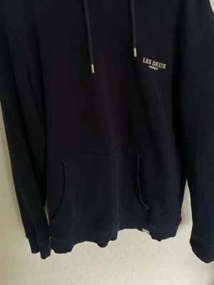 Hoodie från Les Deux  - Mörkblå hoodie från Les Deux med broderad logga på bröstet. Klassisk känguruficka framtill, justerbar huva med snören och ribbade muddar vid ärmslut och nederkant. Perfekt för en avslappnad och stilren look.