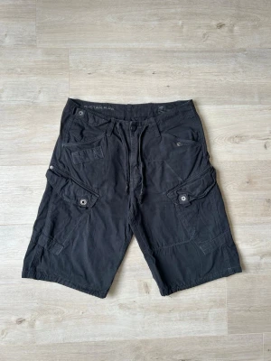 Svarta G-Star RAW Cargoshorts Shorts Y2K Storlek W32 - Mycket fina G-Star shorts i svart färg | Shortsen är i mycket bra skick och sparsamt använda. |  Äkta som allt annat jag säljer! ✅ |  Storlek: W32 | Skick: Mycket Bra Skick - har tyvärr några små slitningar (SE SISTA BILDER) | Färg: Svart | Märke: G-Star RAW | Modell: Shorts |   Hör gärna av dig vid frågor så svarar jag så fort jag kan! 😁 |  (SKU - 178)