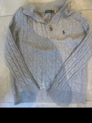 Grå kabelstickad tröja Polo Ralph Lauren - Snygg grå kabelstickad tröja från Polo Ralph Lauren med halv dragkedja och hög krage. Tröjan har ribbade muddar och den klassiska broderade loggan på bröstet. Perfekt för dig som gillar stilrena och tidlösa plagg. Selger fra Norge 