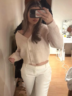 Beige cropped kofta med knappar - Beige cropped kofta i ribbstickat material med stora knappar framtill. Koftan har en djup v-ringning. Den är lite nopprig men inget som syns💕