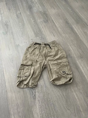 Olivgröna cargoshorts med snörning - Säljer ett par olivgröna cargoshorts med snörning i midjan och stora sidofickor. Shortsen har en avslappnad passform och elastisk midja för extra komfort. Perfekta för varma dagar och enkel att matcha med olika stilar.