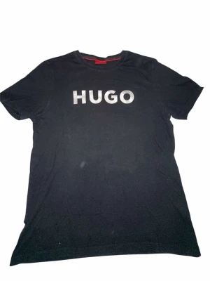 Svart HUGO t-shirt med tryck - Snygg svart t-shirt från HUGO med stort vitt logotryck på bröstet. Klassisk rund halsringning och korta ärmar. Tillverkad i mjuk bomull som känns skön mot huden. Perfekt för en clean och stilren look.