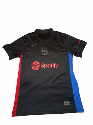 Svart FC Barcelona matchtröja Nike - Snygg svart FC Barcelona matchtröja från Nike med röda och blå sidopaneler. Tröjan har Spotify-logga på bröstet, klubbmärke och Nike-logga. På ryggen finns UNHCR-tryck. Perfekt för dig som älskar fotboll och vill ha en unik supportertröja.