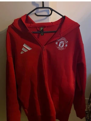 Röd Manchester United hoodie Adidas - Säljer en röd zip-hoodie från Adidas med Manchester United-logga på bröstet och tryck på ryggen. Hoodien har dragkedja framtill, huva och är tillverkad i mjukt material. Perfekt för dig som älskar fotboll och vill ha en sportig stil.
