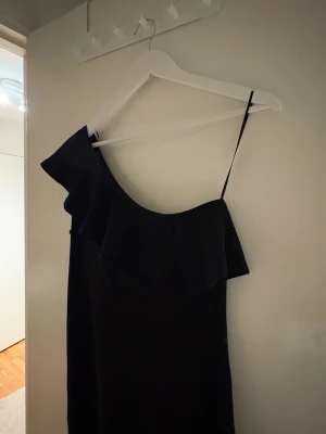 Svart oneshoulder klänning med volang - Säljer en svart oneshoulder klänning med volangdetalj längs urringningen och smalt axelband på ena sidan. Klänningen har en rak passform och är perfekt för dig som vill ha en stilren och trendig look. Materialet känns mjukt och följsamt.