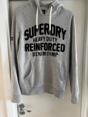 Grå Superdry hoodie - Snygg grå hoodie från Superdry med svart texttryck på bröstet och vita dragsnören i huvan. Passar perfekt nu till de lite svalare sommardagarna skriv gjärna om ni har några frågor 😀👍