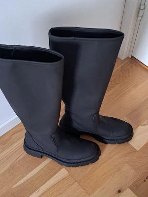 Svarta höga chunky stövlar Zara - Säljer ett par svarta höga stövlar från Zara med chunky sula. Stövlarna har en stilren och minimalistisk design utan dragkedja eller snörning. Tillverkade i ett matt material som ger en modern look. Perfekta för att lyfta vilken outfit som helst.