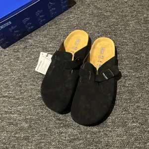 Svarta Birkenstock Boston tofflor - Säljer ett par svarta Birkenstock Boston tofflor i mocka med justerbart spänne och klassisk korksula. Perfekta för dig som vill ha både stil och komfort hemma eller på språng. Innersulan är ljus och formad för extra stöd.