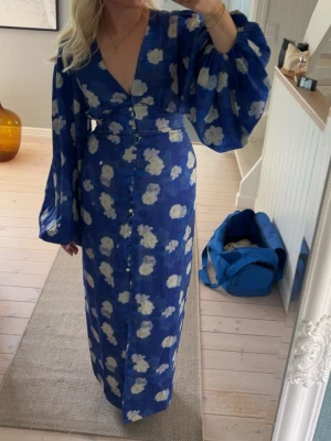 Blommig blå långklänning med v-ringning - Säljer en blå långklänning med vita blommor, v-ringad hals och stora ballongärmar, endast använd 1 gång!Slutsåld på hemsidan 