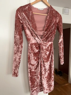Rosa sammetsklänning från NLY One - Supersnygg rosa sammetsklänning från NLY One med omlott och twistad detalj framtill. Klänningen har lång ärm, v-ringning och är i en kort modell. Perfekt för dig som vill sticka ut med en mjuk och glansig look.
