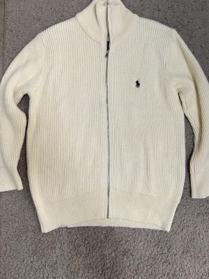 Vit stickad ziptröja från Polo Ralph Lauren - Klassisk vit stickad tröja med dragkedja från Polo Ralph Lauren. Tröjan har hög krage, ribbstickad struktur och den ikoniska mörkblå loggan broderad på bröstet. Perfekt för lager på lager och enkel att matcha med olika outfits.