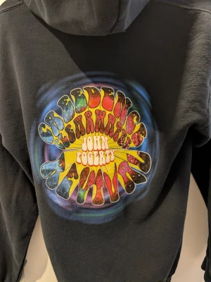 Svart hoodie Creedence Clearwater Revival - Svart hoodie med färgglatt psykedeliskt tryck på ryggen med texten Creedence Clearwater Revival och John Fogerty. Dragkedja framtill och känguruficka. Mjuk bomullsblandning och huva för extra komfort.