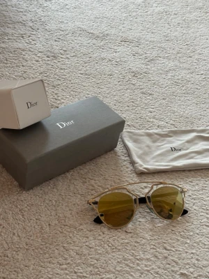 Guldiga Dior solglasögon med gul/grön lins - KVITTO FINNS! Snygga solglasögon från Dior med unika geometriska bågar i guld och gula spegelglas. Glasögonen har svarta skalmar och kommer med originalfodral och ask. Perfekta för dig som vill sticka ut med en trendig accessoar.