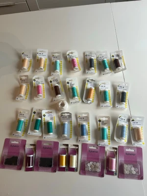 Stort sykit med trådar och tillbehör - Säljer ett komplett sykit med flera rullar sytråd i olika färger som blå, gul, rosa, brun och vit. Inkluderar även sybehör som knappar, tryckknappar och resårband. Perfekt för dig som gillar att sy eller behöver påfyllning till syhörnan.