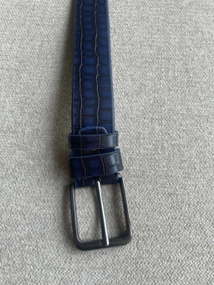 Mörkblått skärp i skinn (krokodil) - Helt nytt snyggt mörkblått bälte i skinn med krokodilpräglad struktur och silverfärgat spänne. Perfekt för att lyfta din outfit med en cool detalj. Bältet har klassisk design och passar till både jeans och kostymbyxor. 120cm. Om du har någon fråga är det bara att höra av sig