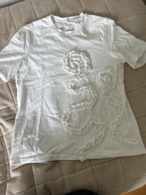Vit t-shirt med volangdetaljer - Säljer en vit t-shirt med korta ärmar och unika volangdetaljer i ett snirkligt mönster på framsidan. T-shirten har en klassisk passform och är tillverkad i mjuk bomull, perfekt för en clean och trendig look.