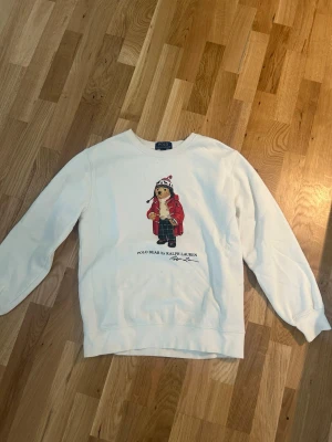 Vit sweatshirt Polo Ralph Lauren - Vit sweatshirt från Polo Ralph Lauren med ikoniskt Polo Bear-tryck framtill. Tröjan har rund halsringning och långa ärmar. Perfekt för dig som gillar klassisk och avslappnad stil med en twist. Storlek L i barn passar XS i herr.