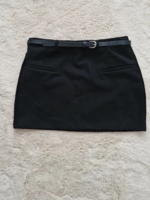 Svart kortkjol med bälte - Stilren svart kortkjol med tillhörande bälte och två diskreta fickor framtill. Kjolen har en klassisk rak passform och är perfekt för att skapa en clean och trendig look.